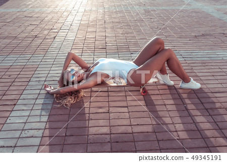 Girl in white bodysuit lying on skateboard in the summer in the city, longboard. Free space for text 49345191