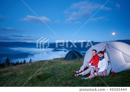 Guy and girl sitting in tent at dawn with view of beautiful landscape 49385444