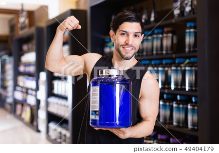 Young bodybuilder showing his biceps and holding pot of sport nutrition products in shop 49404179