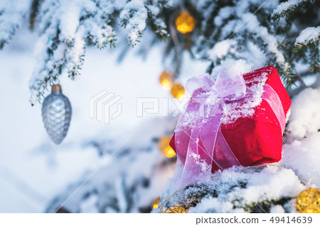 Closeup of a red gift box with a white ribbon next to Christmas decorations on the branches of a Closeup of a red gift box with a white ribbon next to Christmas decorations on the branches of a 49414639