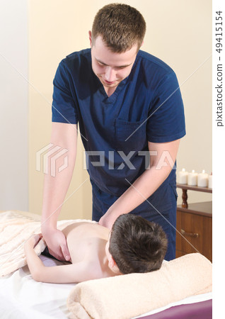 A male physiotherapist masseur makes a healing relaxing massage to a little smiling boy lying on a A male physiotherapist masseur makes a healing relaxing massage to a little smiling boy lying on a 49415154