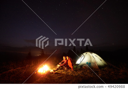 Young couple hikers resting near illuminated tent, camping in mountains at night under starry sky Young couple hikers resting near illuminated tent, camping in mountains at night under starry sky 49420988