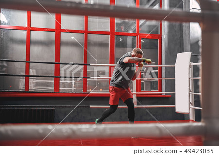 Hard work on boxing ring. Muscular tattooed boxer in sports clothing punching on red ring while Hard work on boxing ring. Muscular tattooed boxer in sports clothing punching on red ring while 49423035