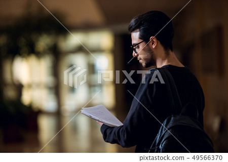 Young student of computer science consults some papers illuminated by the light of a screen in a 49566370
