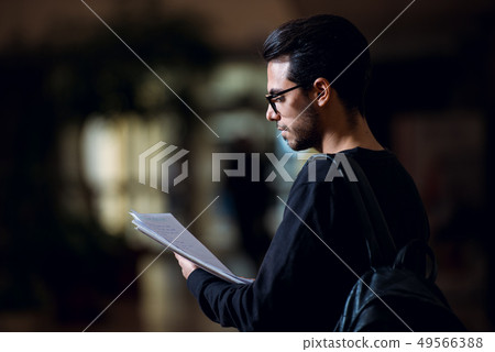 Young student of computer science consults some papers illuminated by the light of a screen in a 49566388