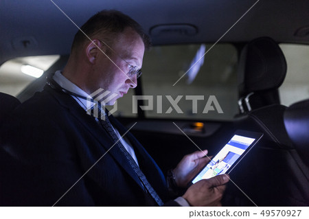 Businessman with a digital tablet sitting in the back seat of a car Businessman with a digital tablet sitting in the back seat of a car 49570927