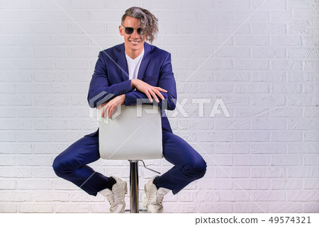 stylish businessman with curly long hair in sunglasses sitting on chair on white background 49574321