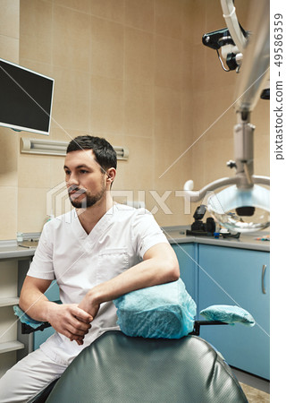 Dentistry for today’s lifestyle. Young dentist looking away while sitting in his cabinet near 49586359
