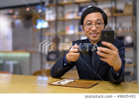 Happy Asian businessman using phone while drinking coffee Happy Asian businessman using phone while drinking coffee 49605493