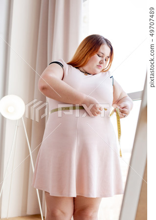 Pleasant plump woman trying to measure her waist Pleasant plump woman trying to measure her waist 49707489