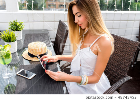 A beautiful woman with long hair is sitting in cafe on summer porch. In hands of tablet, listening A beautiful woman with long hair is sitting in cafe on summer porch. In hands of tablet, listening 49745452