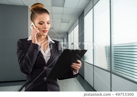 Pretty businesswoman with clipboard talks on phone in modern office 49753300