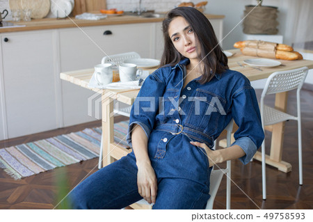 Beautiful woman in denim overalls sits on a chair in the kitchen. Beautiful woman in denim overalls sits on a chair in the kitchen. 49758593