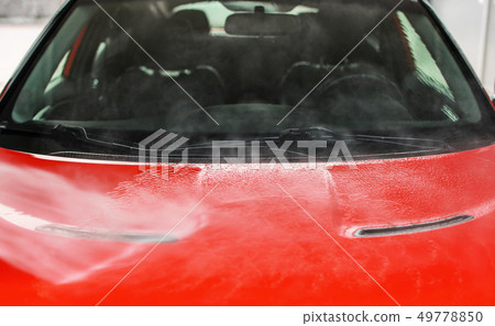 Front hood of red car washed in self service carwash, white water spraying on metal, blurred seats 49778850