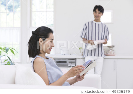 A woman sitting on a couch and reading a book and a man wiping dishes in the kitchen A woman sitting on a couch and reading a book and a man wiping dishes in the kitchen 49836179