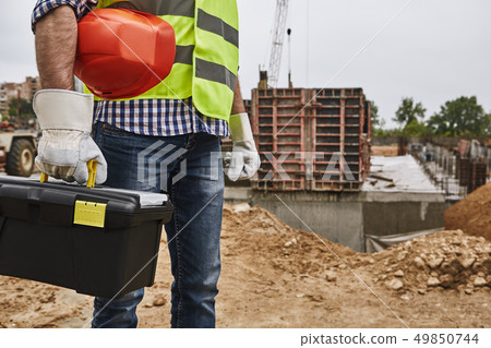 Ready to work! Cropped image of a builder in working uniform holding red helmet and carrying toolbox 49850744