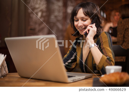 Beautiful businesswoman working on her laptop while having a conversation on the phone in a coffee 49866629