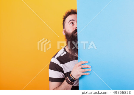 Cool bearded man looking up behind a blue panel over yellow background. 49866838