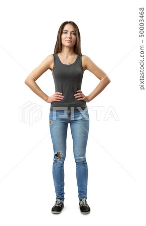 Front view of young attractive woman in gray sleeveless top and blue jeans standing with arms on 50003468