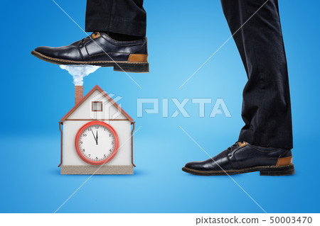 Side crop close-up of man's legs, one foot raised to step on a little house with a big clock on the 50003470