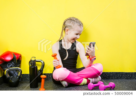 Young child girl with a mobile phone sitting on the floor near the dumbbells, boxing gloves and a 50009004