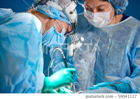 Surgeon performing cosmetic surgery in hospital operating room. Surgeon in mask wearing loupes 50041117