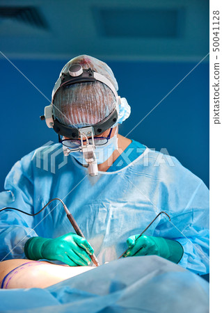 Surgeon performing cosmetic surgery in hospital operating room. Surgeon in mask wearing loupes 50041128