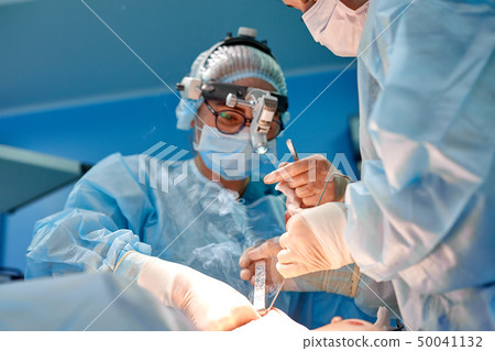 Surgeon performing cosmetic surgery in hospital operating room. Surgeon in mask wearing loupes Surgeon performing cosmetic surgery in hospital operating room. Surgeon in mask wearing loupes 50041132