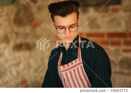 Young male artist in aprong and glasses stand in front of brick wall 50063772
