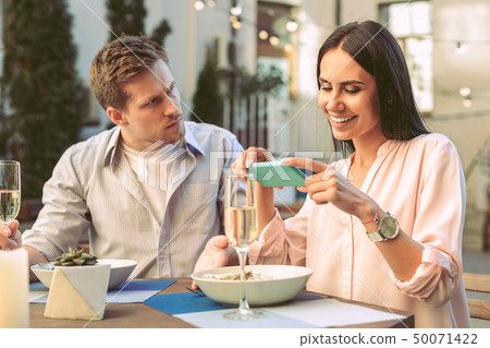 Beaming dark-haired woman in light blouse photographing her dinner Beaming dark-haired woman in light blouse photographing her dinner 50071422