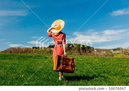 Beautiful girl in red dress and hat with suitcase on meadow Beautiful girl in red dress and hat with suitcase on meadow 50076847