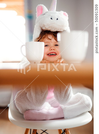 A portrait of small girl in unicorn mask sitting at the table at home. A portrait of small girl in unicorn mask sitting at the table at home. 50091309
