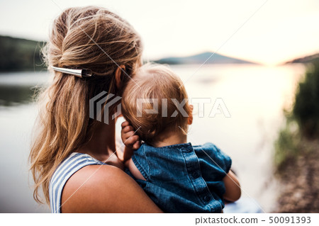 A rear view of mother with a toddler daughter outdoors by the river in summer. A rear view of mother with a toddler daughter outdoors by the river in summer. 50091393