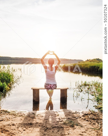 The girl enjoys a beautiful sunset sits on a bench by the river and shows hands fingers heart sign The girl enjoys a beautiful sunset sits on a bench by the river and shows hands fingers heart sign 50107324