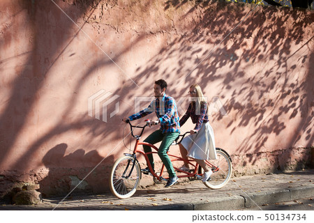 Tourist couple, bearded man and blond woman on tandem bicycle on background of red crackled wall. 50134734