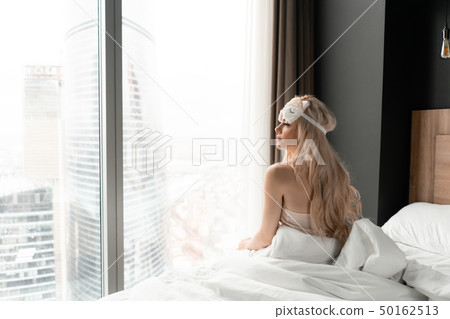 Morning in hotel room. Young woman sits on comfortable bed in mask for sleeping on head. Window of a 50162513