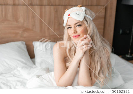 Morning in hotel room. Young woman sits on comfortable bed in mask for sleeping on head. White Morning in hotel room. Young woman sits on comfortable bed in mask for sleeping on head. White 50162551