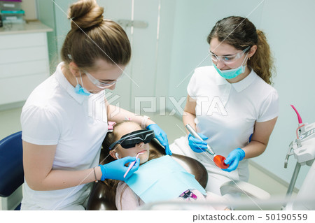 Little girl in VR glasses and wireless headphones sitting in dental chair. Dentist with assistant Little girl in VR glasses and wireless headphones sitting in dental chair. Dentist with assistant 50190559