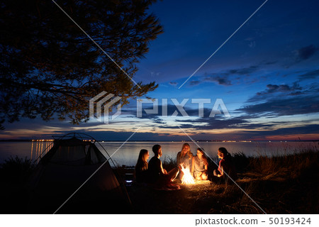 Night summer camping on shore. Group of young tourists around campfire near tent under evening sky Night summer camping on shore. Group of young tourists around campfire near tent under evening sky 50193424