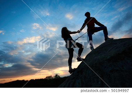 woman with rope engaged in sports of rock climbing on the top of cliff. Her friend helping her 50194997