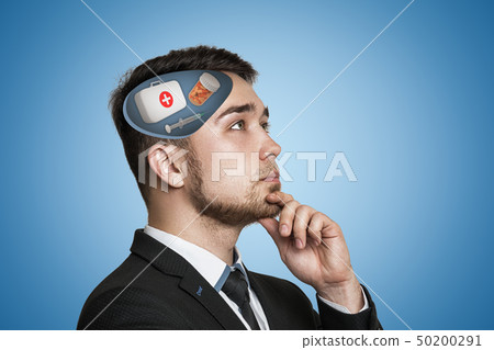 Young businessman thinking with first aid medical box, pills jar and syringe in his head on blue Young businessman thinking with first aid medical box, pills jar and syringe in his head on blue 50200291