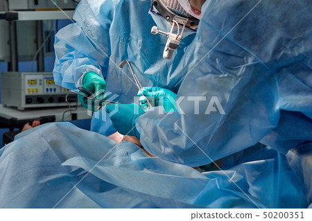 Surgeon performing cosmetic surgery in hospital operating room. Surgeon in mask wearing loupes 50200351