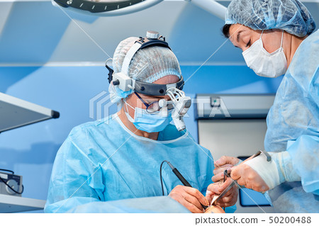 Surgeon performing cosmetic surgery in hospital operating room. Surgeon in mask wearing loupes Surgeon performing cosmetic surgery in hospital operating room. Surgeon in mask wearing loupes 50200488
