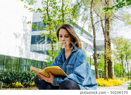 Young student girl in denim jacket reading a book at alley near modern building of University. 50206570