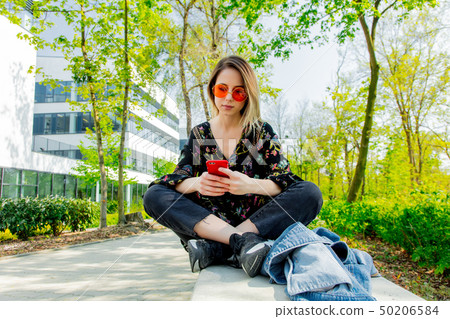 Young woman in sunglasses with mobile phone sitting on green alley near modern building of Young woman in sunglasses with mobile phone sitting on green alley near modern building of 50206584