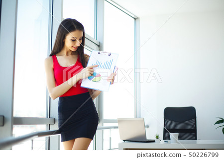 Office. Business Woman Standing at the Grand Window. Light Modern Office. Dressed in Red Sweater and Office. Business Woman Standing at the Grand Window. Light Modern Office. Dressed in Red Sweater and 50226799