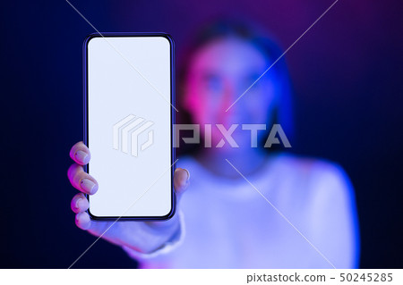 Girl showing blank smartphone screen over neon studio background Girl showing blank smartphone screen over neon studio background 50245285