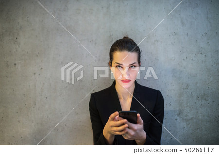 A front view of young business woman with smartphone standing against concrete wall in office. A front view of young business woman with smartphone standing against concrete wall in office. 50266117