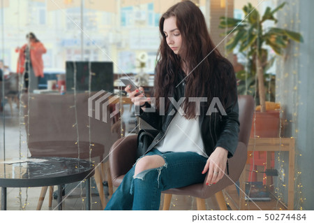 Concentrated hipster girl looking at smartphone during online video call, sitting in coffee shop 50274484