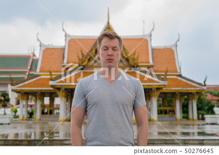 Young tourist man with eyes closed against view of the Buddhist temple in Bangkok Young tourist man with eyes closed against view of the Buddhist temple in Bangkok 50276645